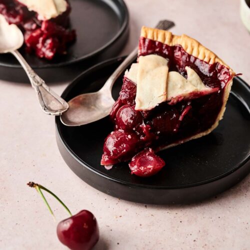 Two slices of Cherry Pie with lattice crust sit on black plates, each accompanied by a spoon and a fresh cherry, all arranged on a light pink surface.