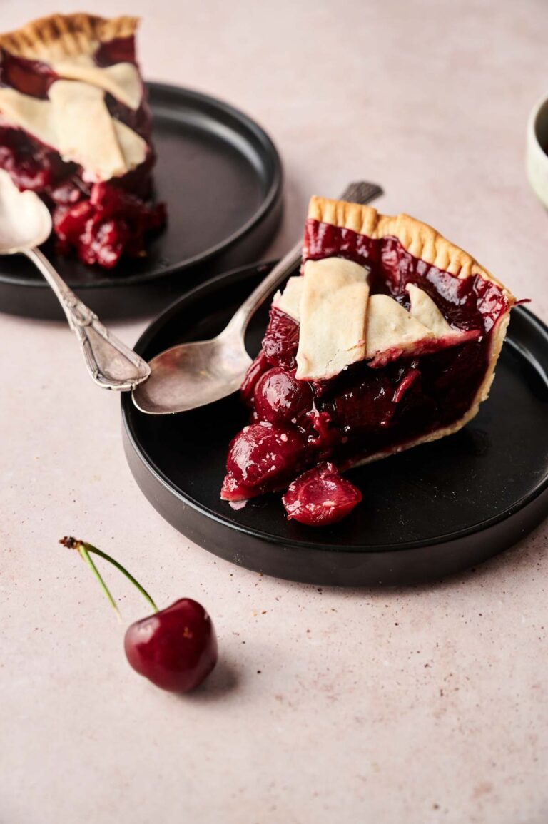 Two slices of Cherry Pie with lattice crust sit on black plates, each accompanied by a spoon and a fresh cherry, all arranged on a light pink surface.