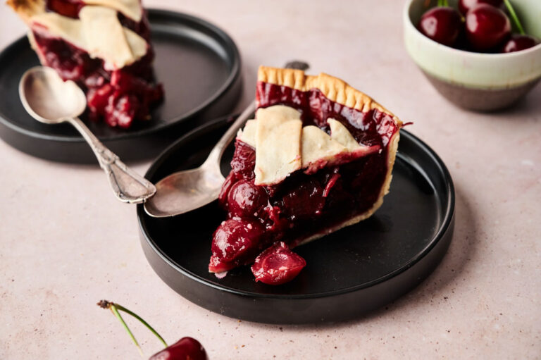 A slice of cherry pie with a golden lattice crust sits on a black plate with a spoon; another cherry pie slice and a bowl of cherries are in the background.