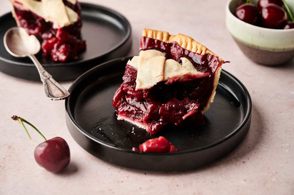 A slice of cherry pie with lattice crust sits on a black plate, a fresh cherry beside it, showcasing the irresistible appeal of classic Cherry Pie with more slices and cherries in the background.