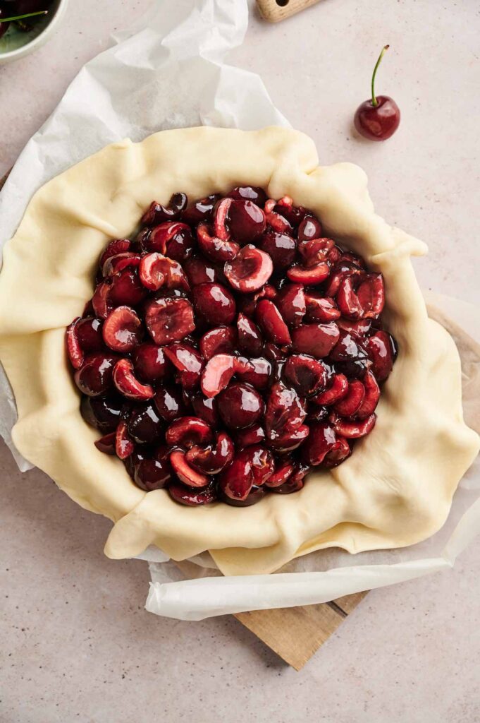 Unbaked cherry pie with fresh cherry filling in a pie crust, dough edges draped over the dish, rests on a light surface&mdash;a classic Cherry Pie scene, complete with a single cherry nearby.