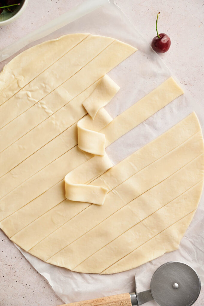A round sheet of pastry dough is cut into strips, with some folded over each other next to a pizza cutter and a cherry&mdash;perfect for creating a classic Cherry Pie lattice&mdash;on a light surface.