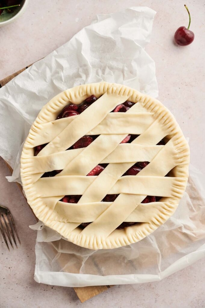 A Cherry Pie with a lattice crust sits unbaked on parchment paper, ready for the oven. A cherry and forks rest nearby on the light surface, inviting a taste.