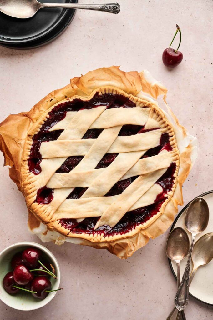 A classic Cherry Pie with a golden lattice crust sits on parchment paper, surrounded by fresh cherries, plates, and spoons on a light-colored surface.