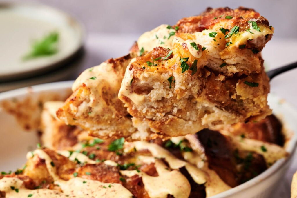 A close-up of a serving of bread pudding, reminiscent of an Eggs Benedict Casserole, topped with creamy sauce and garnished with chopped herbs, being lifted from a baking dish.