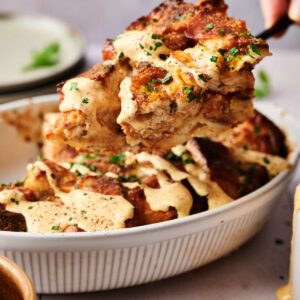 A close-up of bread pudding being served from a white baking dish, topped with a creamy sauce and chopped herbs, reminiscent of a savory Eggs Benedict Casserole.