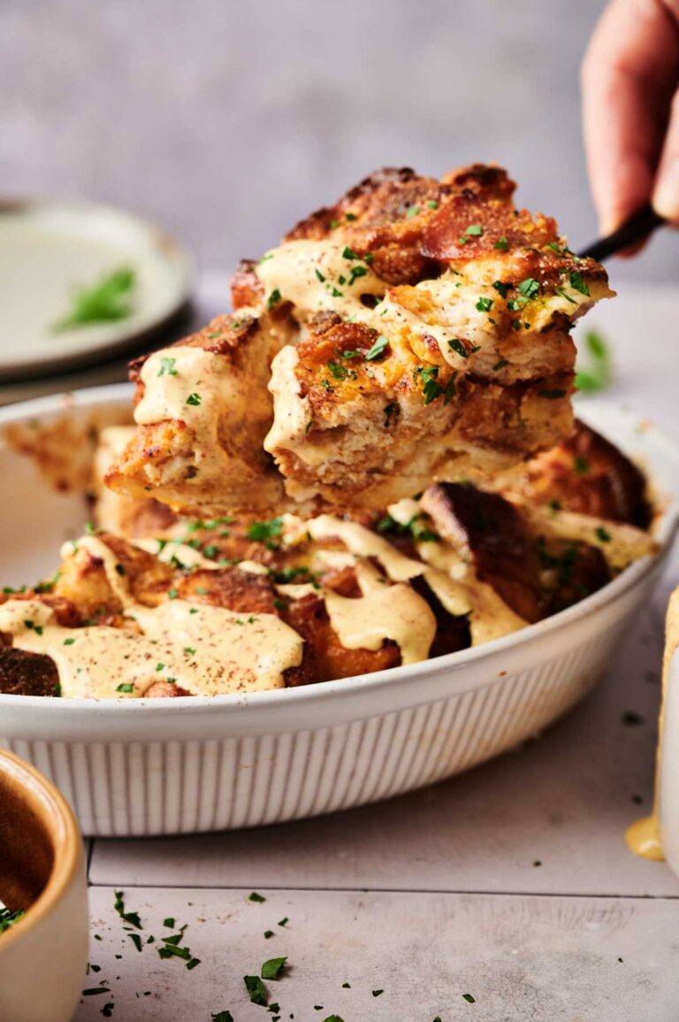 A close-up of bread pudding being served from a white baking dish, topped with a creamy sauce and chopped herbs, reminiscent of a savory Eggs Benedict Casserole.