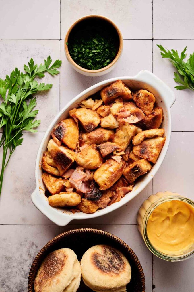 Oval dish with browned bread pieces and ham, surrounded by fresh parsley, a small bowl of chopped herbs, a jar of yellow sauce, and two toasted English muffins&mdash;perfect for serving as an Eggs Benedict Casserole.