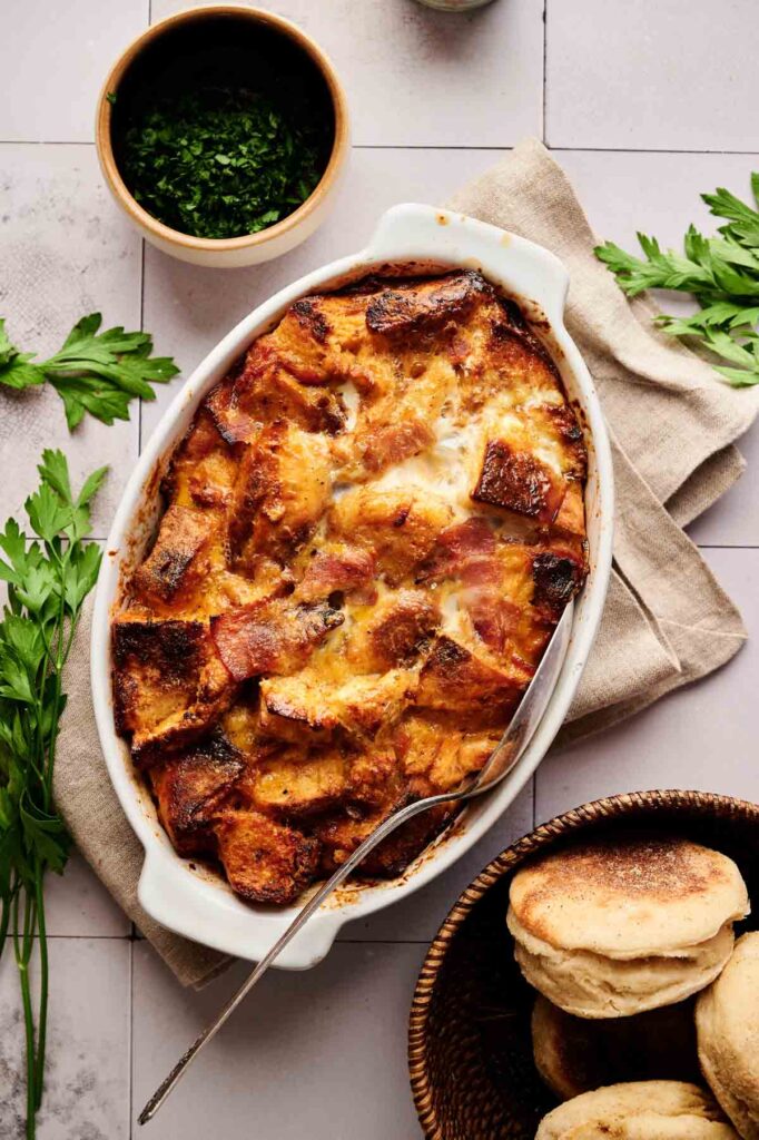 An oval baking dish filled with golden-brown Eggs Benedict Casserole strata, surrounded by fresh parsley, a small bowl of chopped herbs, and a basket of English muffins.