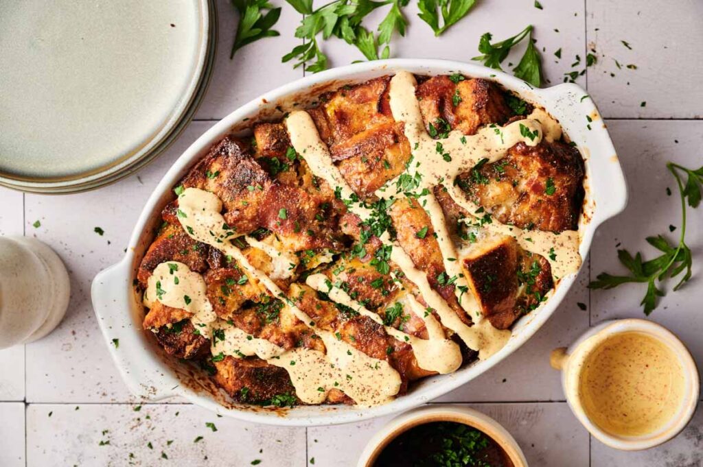 An oval baking dish filled with savory Eggs Benedict Casserole, topped with creamy sauce and chopped parsley, surrounded by plates, a cup of sauce, and fresh herbs on a tiled surface.