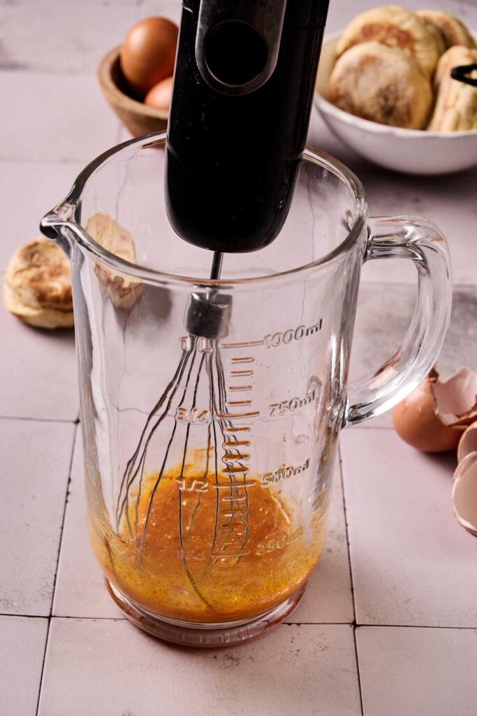 A hand mixer blends eggs in a glass measuring jug; eggshells and a bowl with food for an Eggs Benedict Casserole are visible in the background.