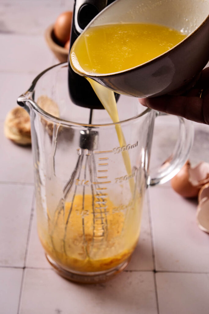 Melted butter is being poured from a bowl into a glass measuring cup containing beaten eggs and a whisk, as eggshells and eggs sit nearby&mdash;ideal prep for an Eggs Benedict Casserole.