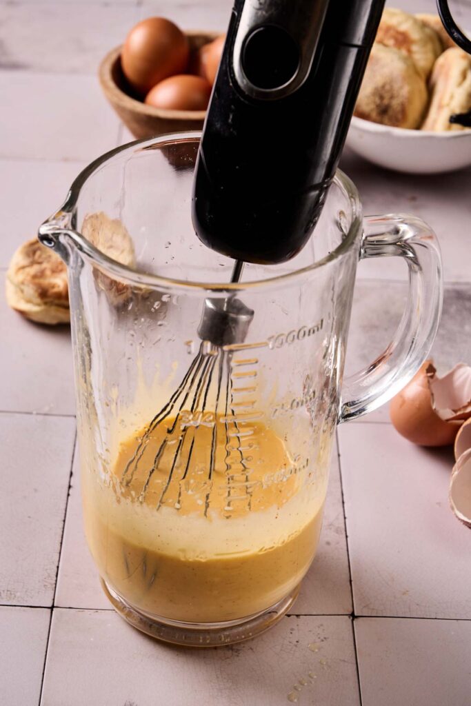 A hand mixer whisking eggs in a glass measuring cup, with ingredients for Eggs Benedict Casserole and baked goods in the background.