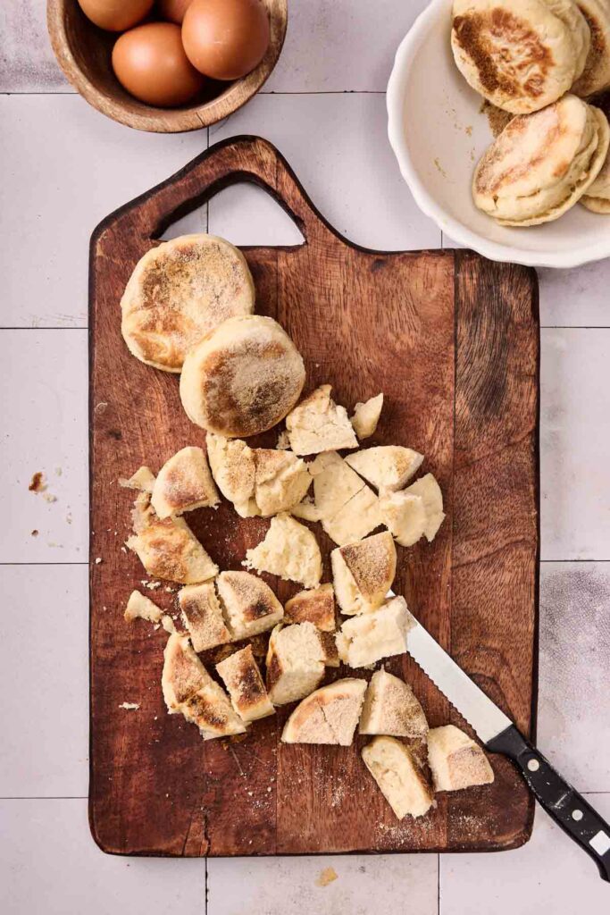 A wooden cutting board with a partially sliced English muffin, a knife, whole English muffins on a plate, and a bowl of eggs nearby&mdash;perfect for preparing Eggs Benedict Casserole.