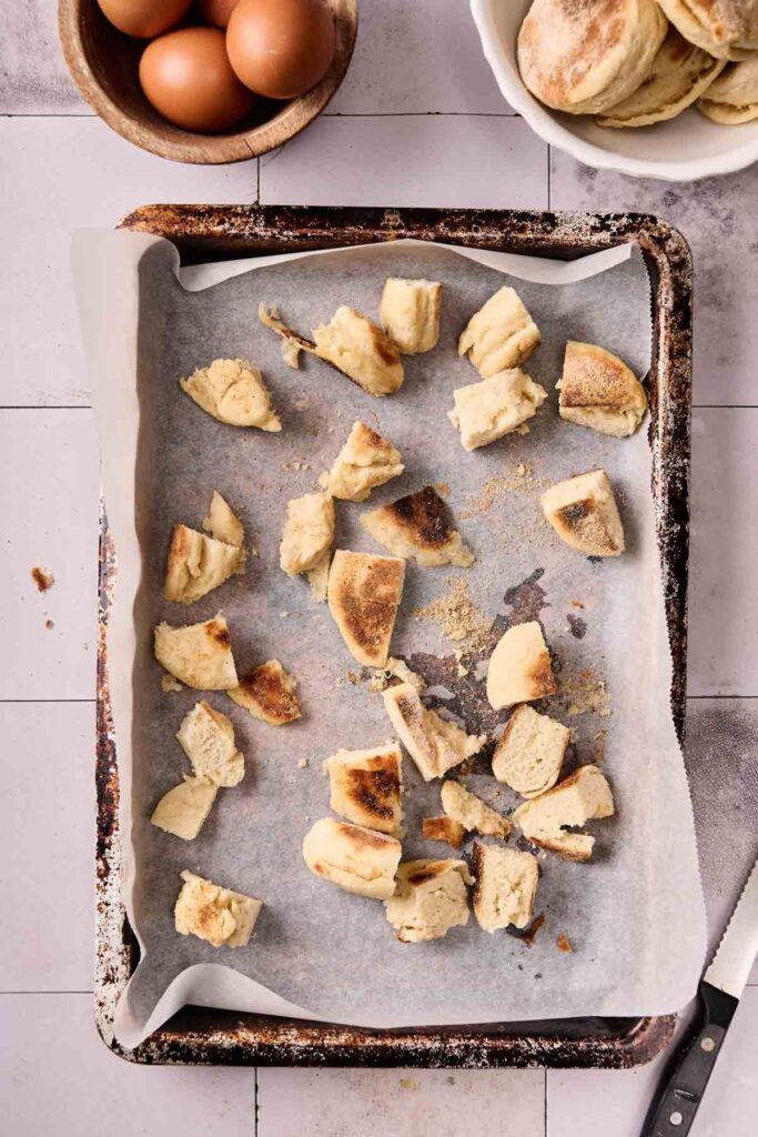 Baking sheet with parchment paper holds pieces of torn biscuit dough for an Eggs Benedict Casserole. Bowl of brown eggs and plate of baked biscuits are nearby. Knife rests on the side.