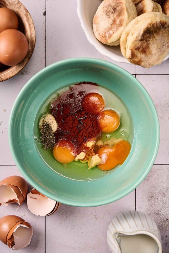 A green bowl with cracked eggs, spices, and seasoning, surrounded by whole eggs, egg shells, biscuits, and a small pitcher on a white tile surface&mdash;perfect for prepping an Eggs Benedict Casserole.