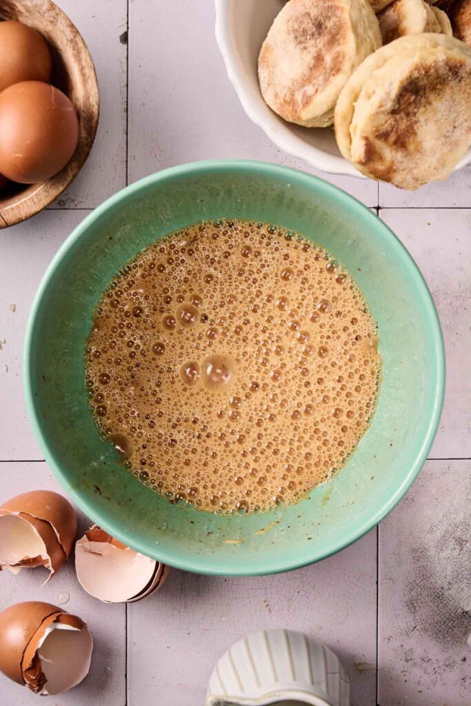 A bowl of beaten eggs sits on a tiled surface, surrounded by eggshells, whole eggs in a wooden bowl, a white bowl of English muffins&mdash;perfect ingredients for making an Eggs Benedict Casserole&mdash;and a small ceramic container.