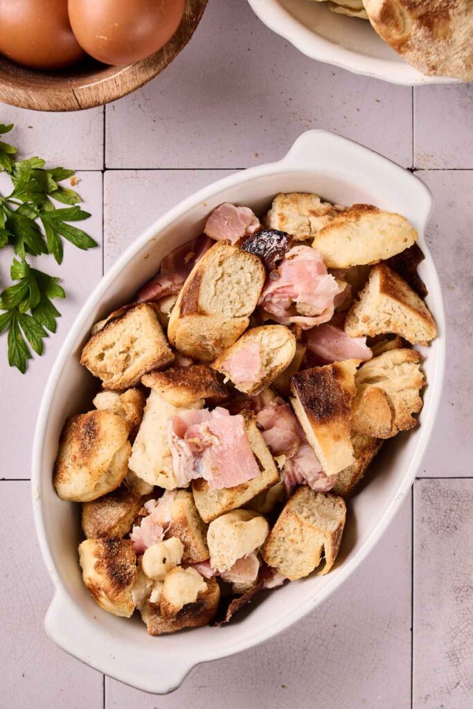 An oval white dish filled with chunks of bread and ham, resembling an Eggs Benedict Casserole, sits on a tiled surface next to a sprig of parsley and a bowl of eggs.