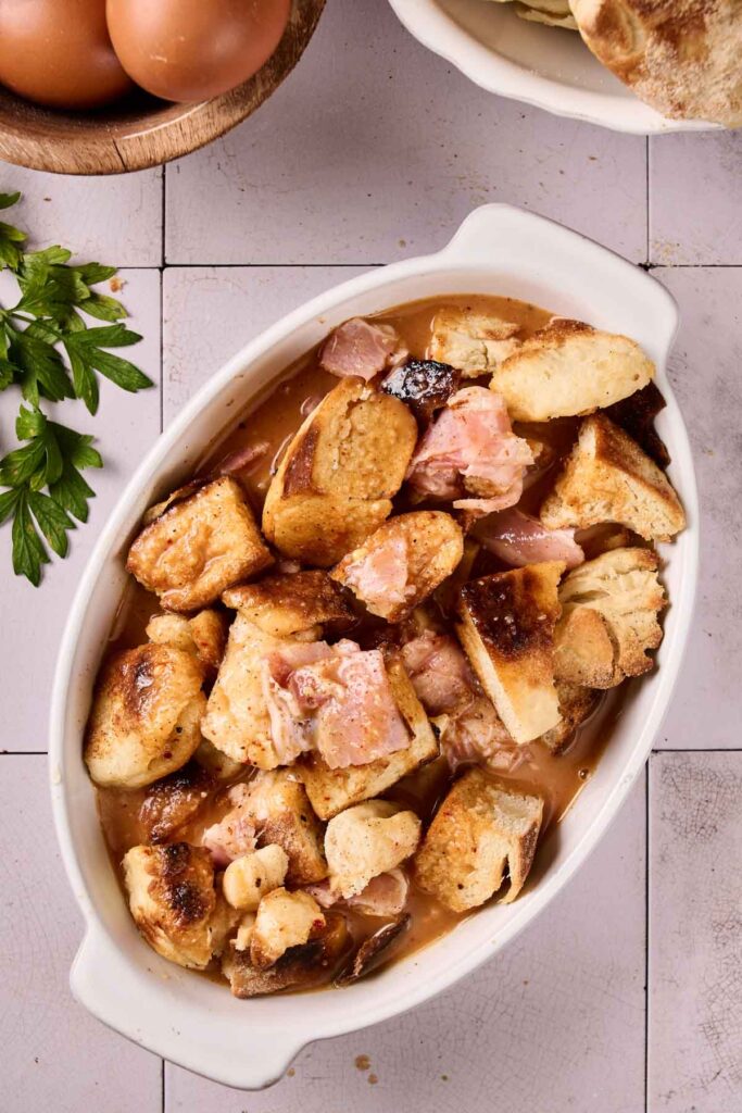 Oval dish filled with chunks of bread and pieces of ham in a brown broth, shown on a tiled surface with parsley and Eggs Benedict Casserole ingredients like eggs nearby.