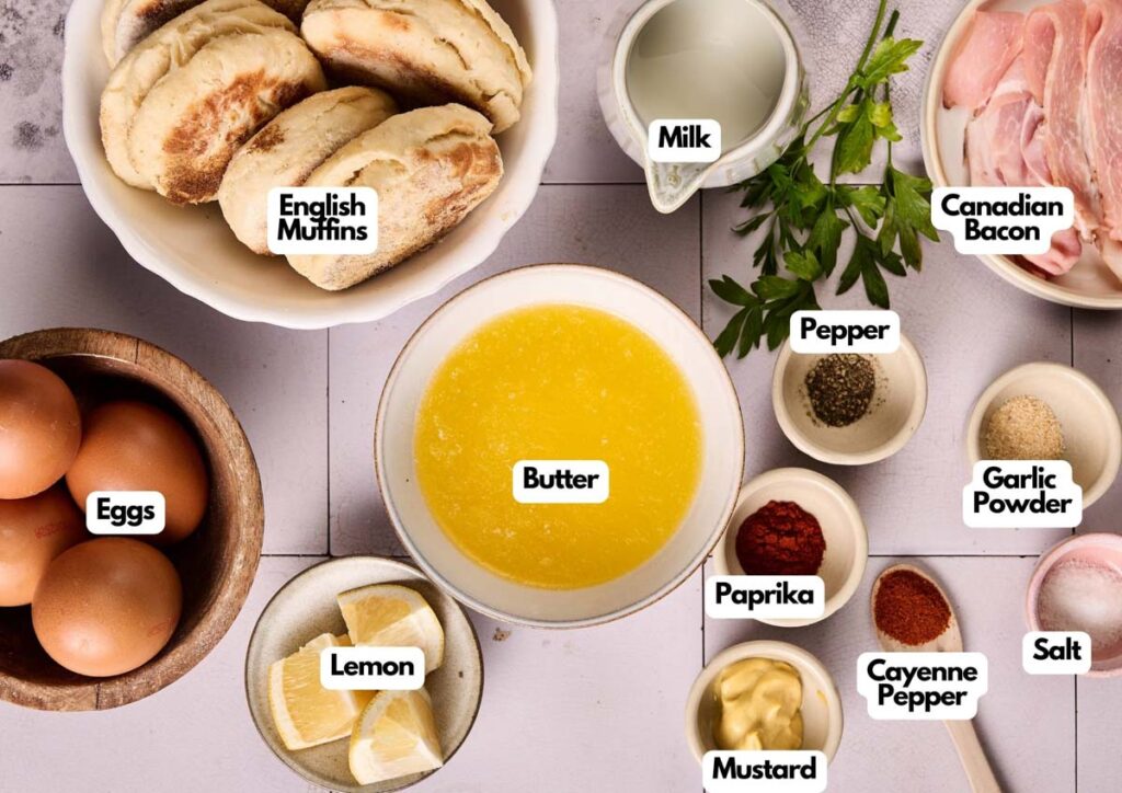 Top-down view of Eggs Benedict Casserole ingredients, including English muffins, eggs, lemon, butter, milk, Canadian bacon, parsley, pepper, paprika, mustard, cayenne, garlic powder, and salt.