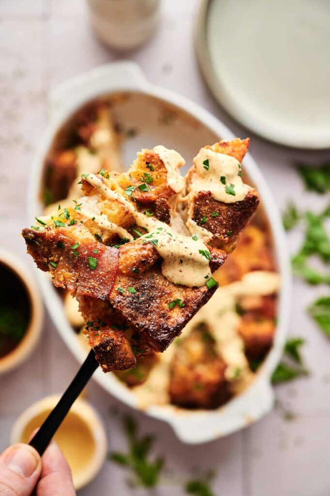 A close-up of a fork holding a piece of baked bread pudding topped with creamy sauce and chopped herbs, with a dish of Eggs Benedict Casserole in the background.