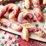 Twisted Candy Cane Cookies dusted with powdered sugar, surrounded by festive red, white, and green holiday sprinkles on a vibrant red surface.