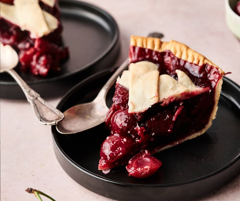 A slice of cherry pie with a golden lattice crust sits on a black plate, a spoon beside it. Another plate with cherry pie is visible in the background, making this a tempting scene for dessert lovers.