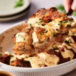 A close-up of a serving spoon lifting a portion of savory Eggs Benedict Casserole bread pudding, topped with creamy sauce and chopped herbs, from a white casserole dish.