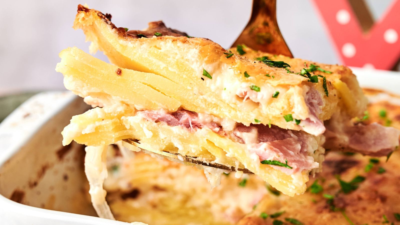 A close-up of a serving of potato gratin with visible layers of sliced potatoes, ham, and creamy sauce, being lifted from a baking dish.