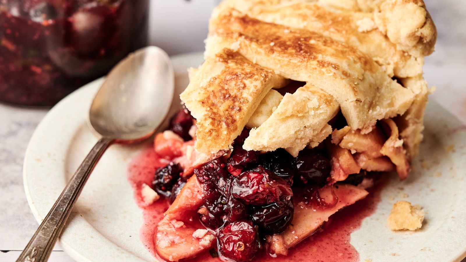 A slice of mixed berry pie with a golden crust sits on a plate next to a spoon, with fruit filling spilling out.