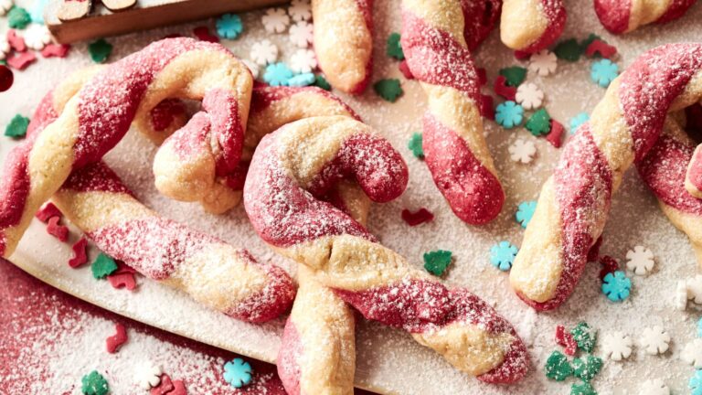 Candy cane-shaped cookies with red and white twisted dough, dusted with powdered sugar, surrounded by festive sprinkles in the shapes of snowflakes and holly leaves.