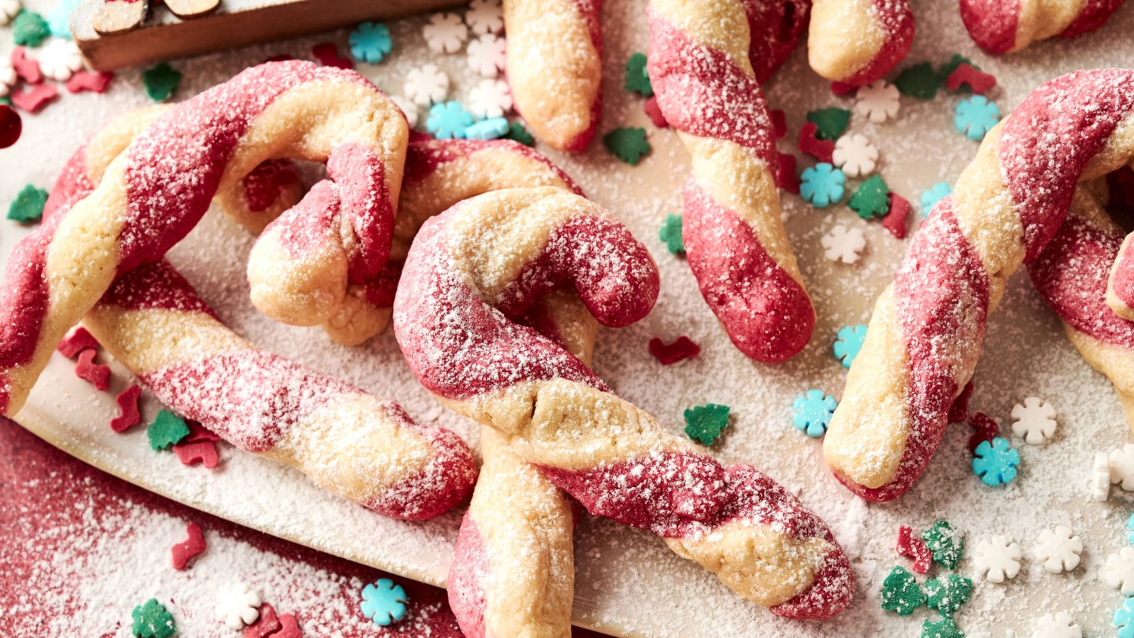 Twisted red and white candy cane cookies dusted with powdered sugar, surrounded by decorative snowflake and tree-shaped sprinkles.