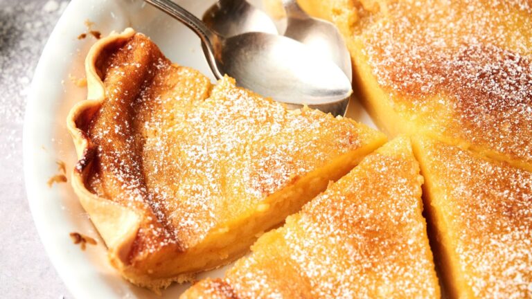 A close-up of a sliced lemon tart dusted with powdered sugar, served on a white plate with two metal spoons.