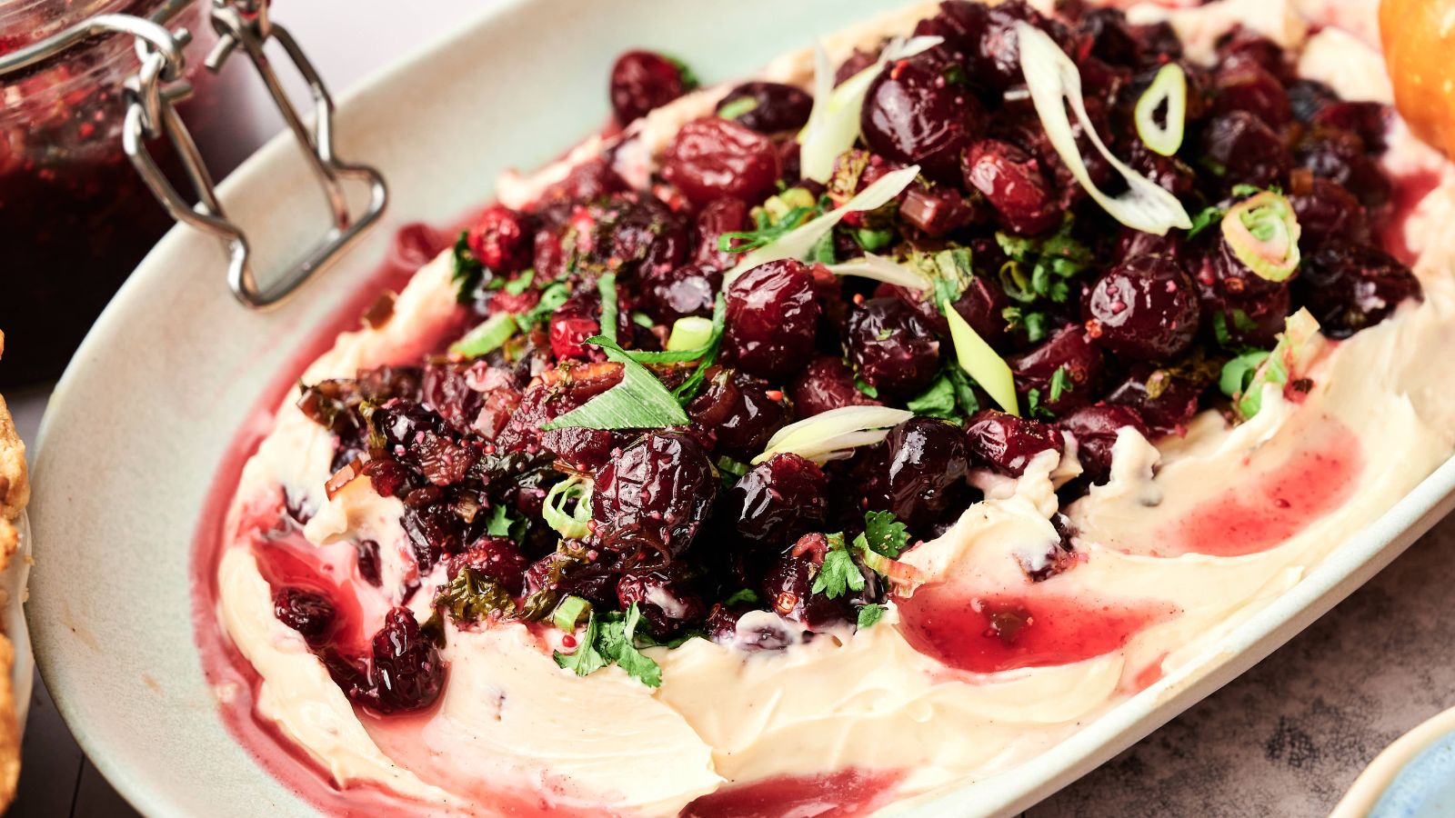A platter of whipped cheese topped with roasted cranberries, chopped herbs, and sliced green onions.