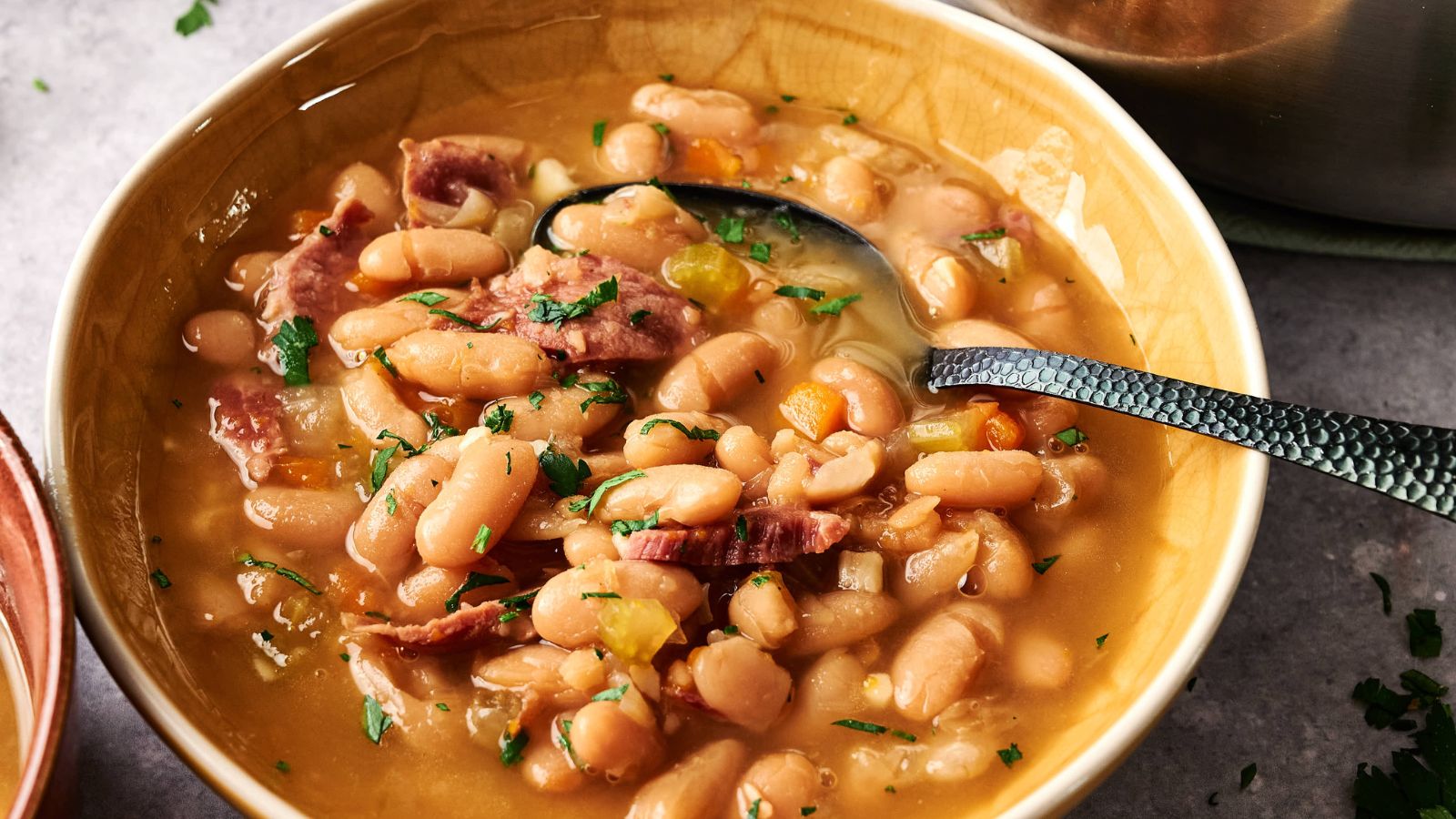 A bowl of white bean soup with pieces of ham, garnished with chopped parsley, and a spoon resting in the bowl.