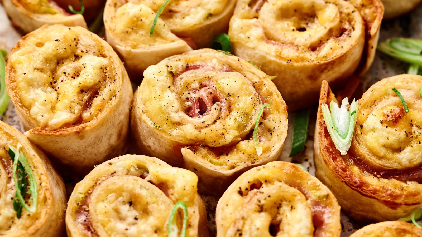 Close-up of baked pinwheel appetizers filled with cheese and ham, topped with black pepper and garnished with sliced green onions.