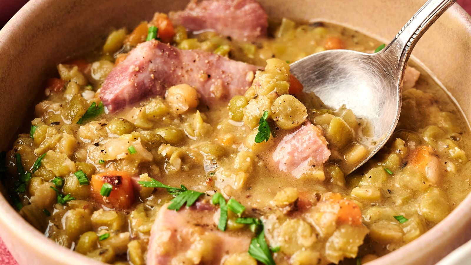 A bowl of split pea soup with chunks of ham, diced carrots, and herbs, with a spoon resting inside.