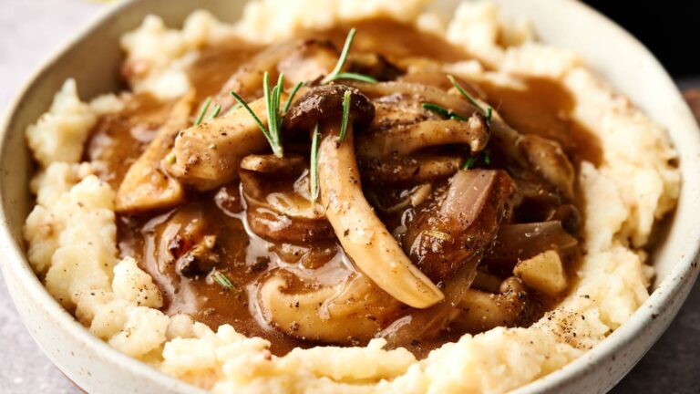 A bowl of mashed potatoes topped with mushroom and onion gravy, garnished with fresh rosemary.