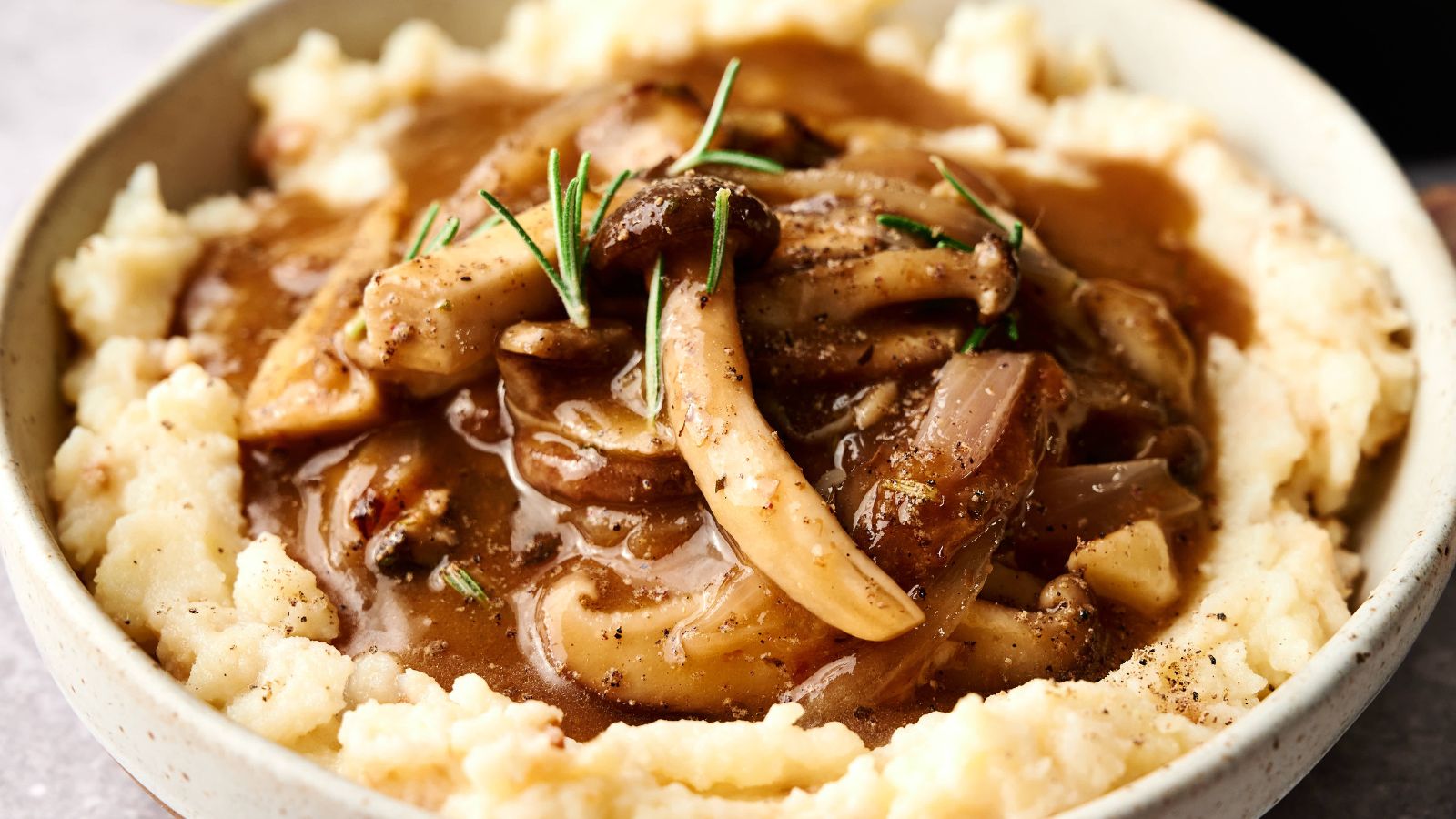 A bowl of mashed potatoes topped with mushroom and onion gravy, garnished with rosemary.