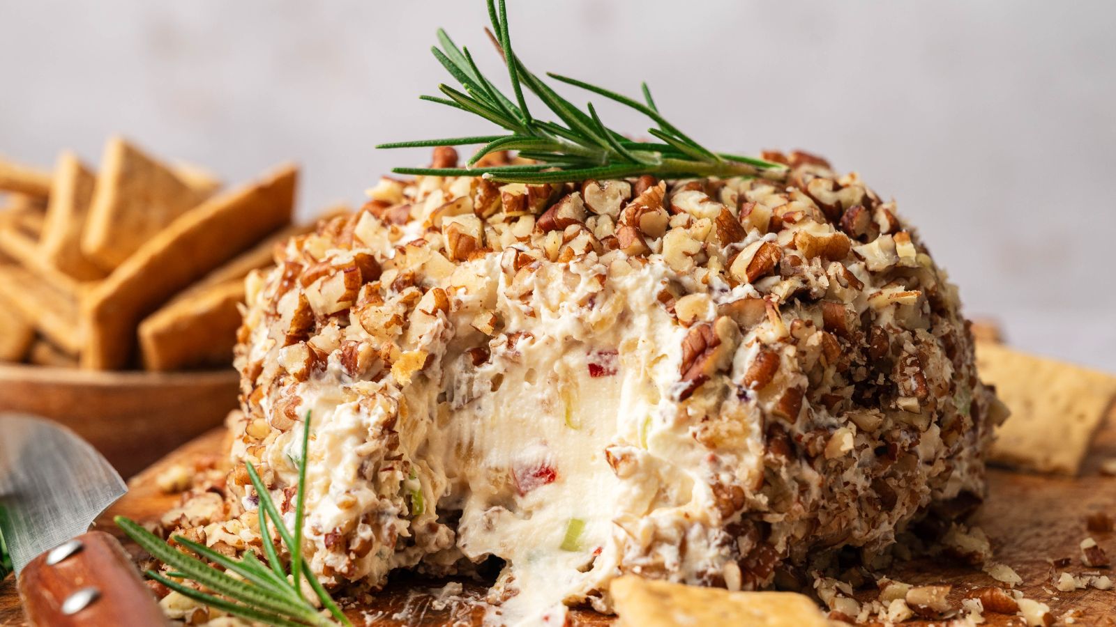 A round cheese ball coated in chopped nuts, garnished with a sprig of rosemary, sits on a wooden board with crackers beside it; a portion is scooped out.