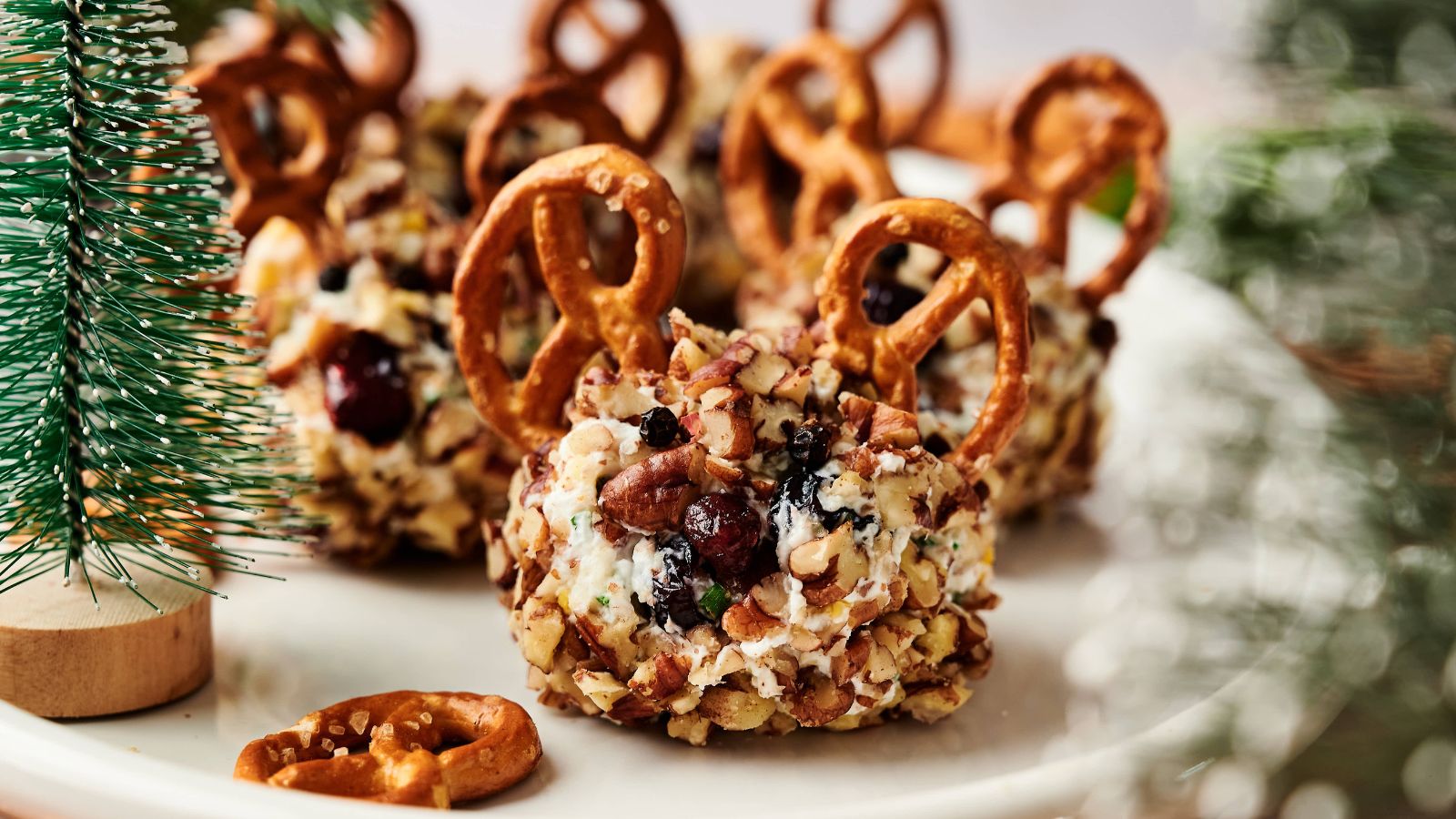 Cheese balls coated with chopped nuts, topped with cranberries and pretzels, arranged on a white plate near a small artificial pine tree.