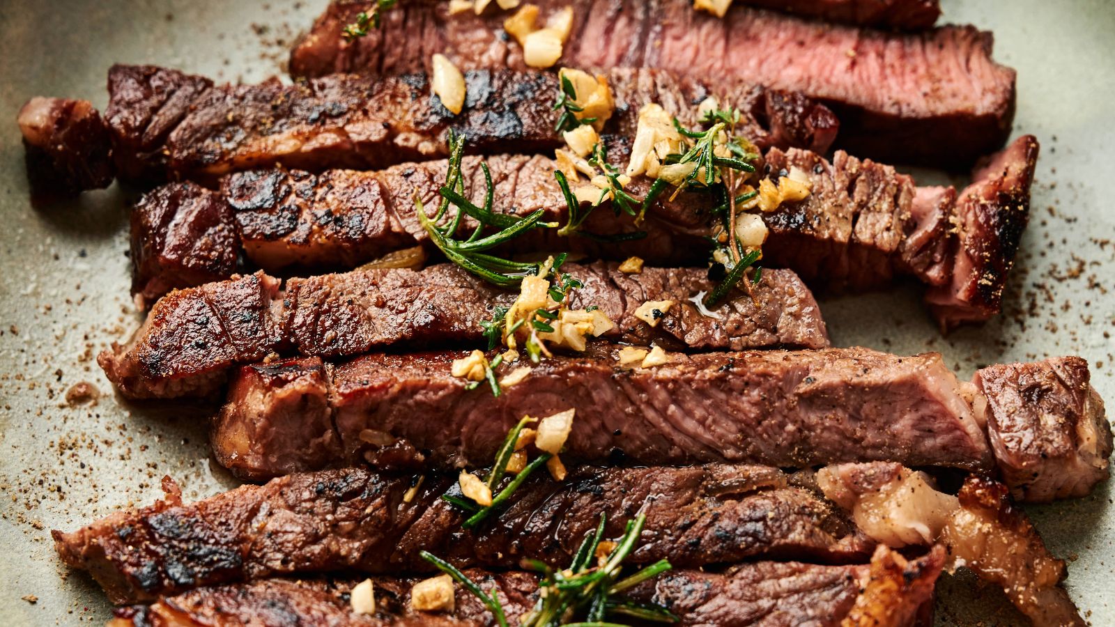 Sliced grilled steak topped with chopped garlic and sprigs of fresh rosemary on a plate.