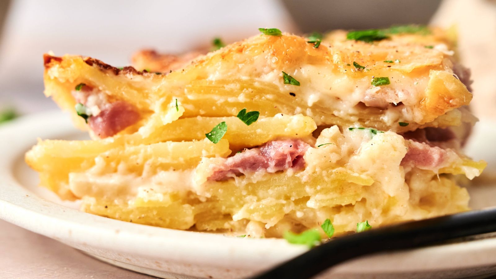 A close-up of a slice of potato casserole with layers of potatoes, ham, and creamy sauce, garnished with chopped parsley on a plate.