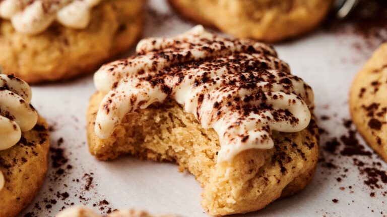 A close-up of a cookie with white frosting and cocoa powder, showing a bite taken out, placed on a parchment-lined surface with other cookies in the background.