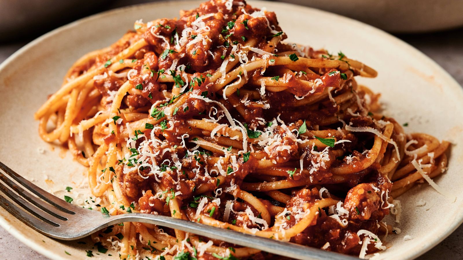 A plate of spaghetti topped with meat sauce, grated cheese, and chopped herbs, with a fork resting on the plate.