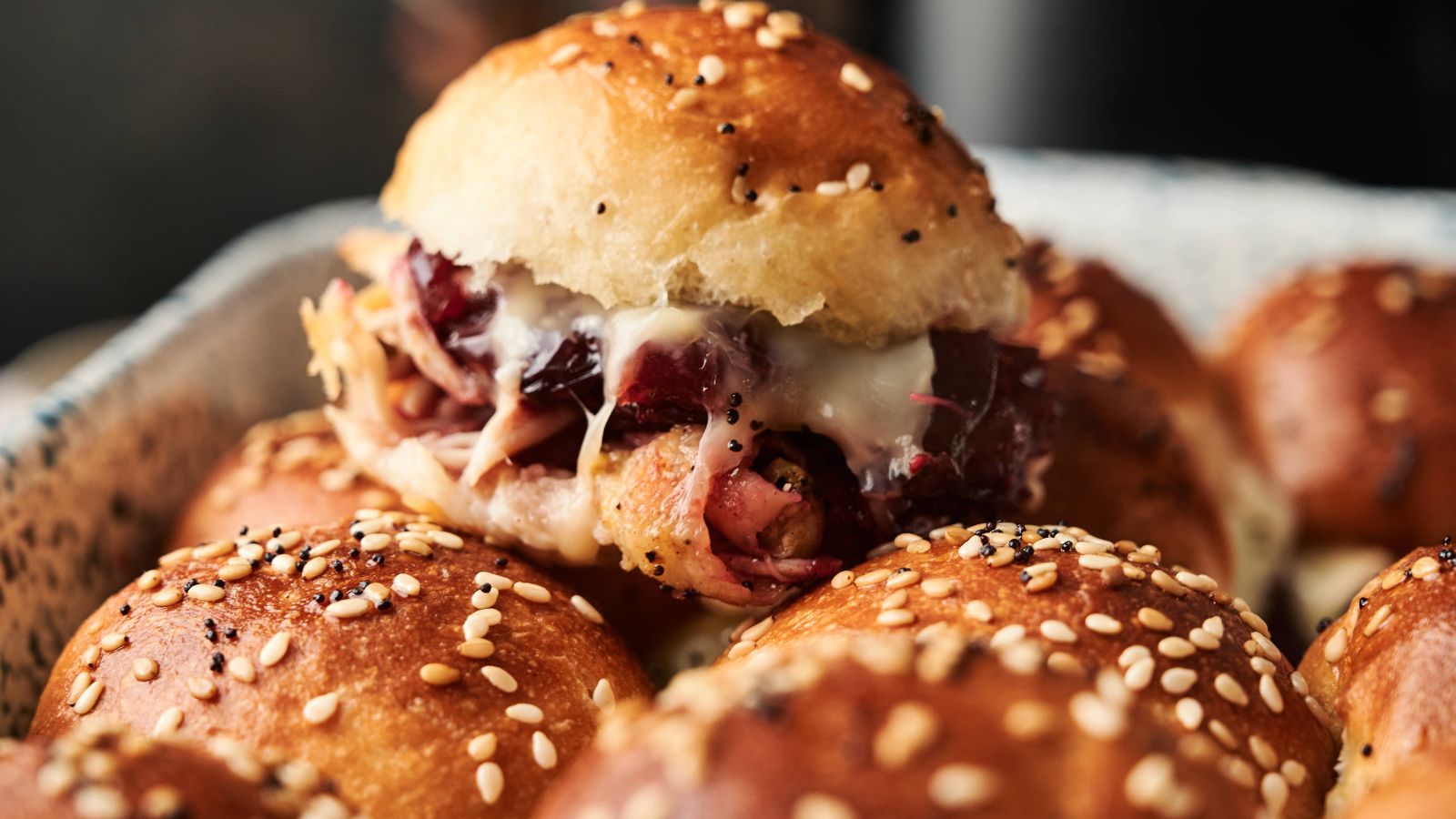 A close-up of a tray of sesame seed slider buns filled with shredded meat, melted cheese, and cranberry sauce.