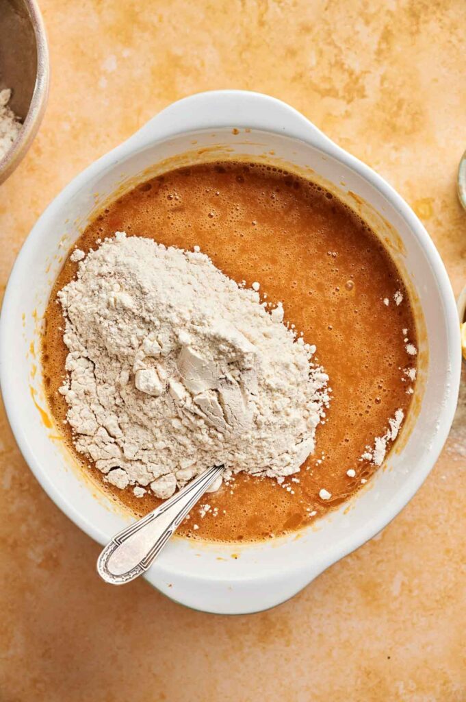 A mixing bowl with brown pumpkin cake batter and a mound of flour on top, with a metal spoon resting inside, sits on a light-colored countertop.