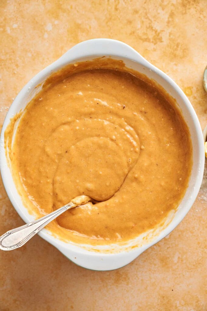 A white bowl filled with thick, tan-brown pumpkin cake batter and a spoon resting in it, placed on a light brown surface.