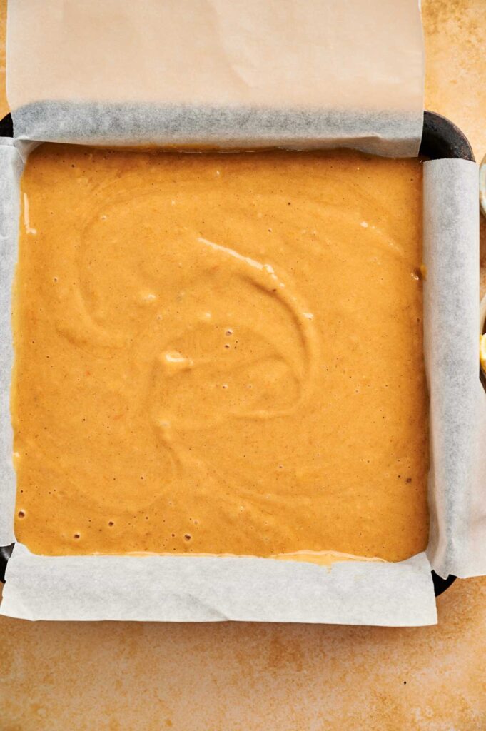 A square baking pan lined with parchment paper is filled with an even layer of light brown pumpkin cake batter, ready to be baked.