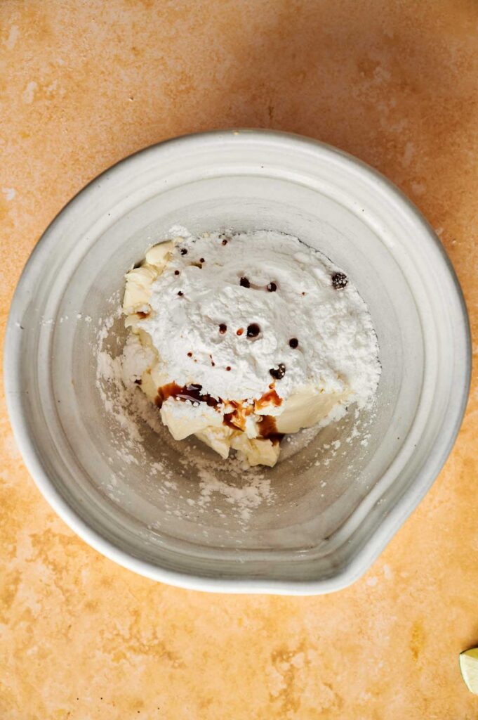 A white mixing bowl containing cream cheese, powdered sugar, vanilla extract, and a few brown specks for making Pumpkin Cake, on a beige countertop.